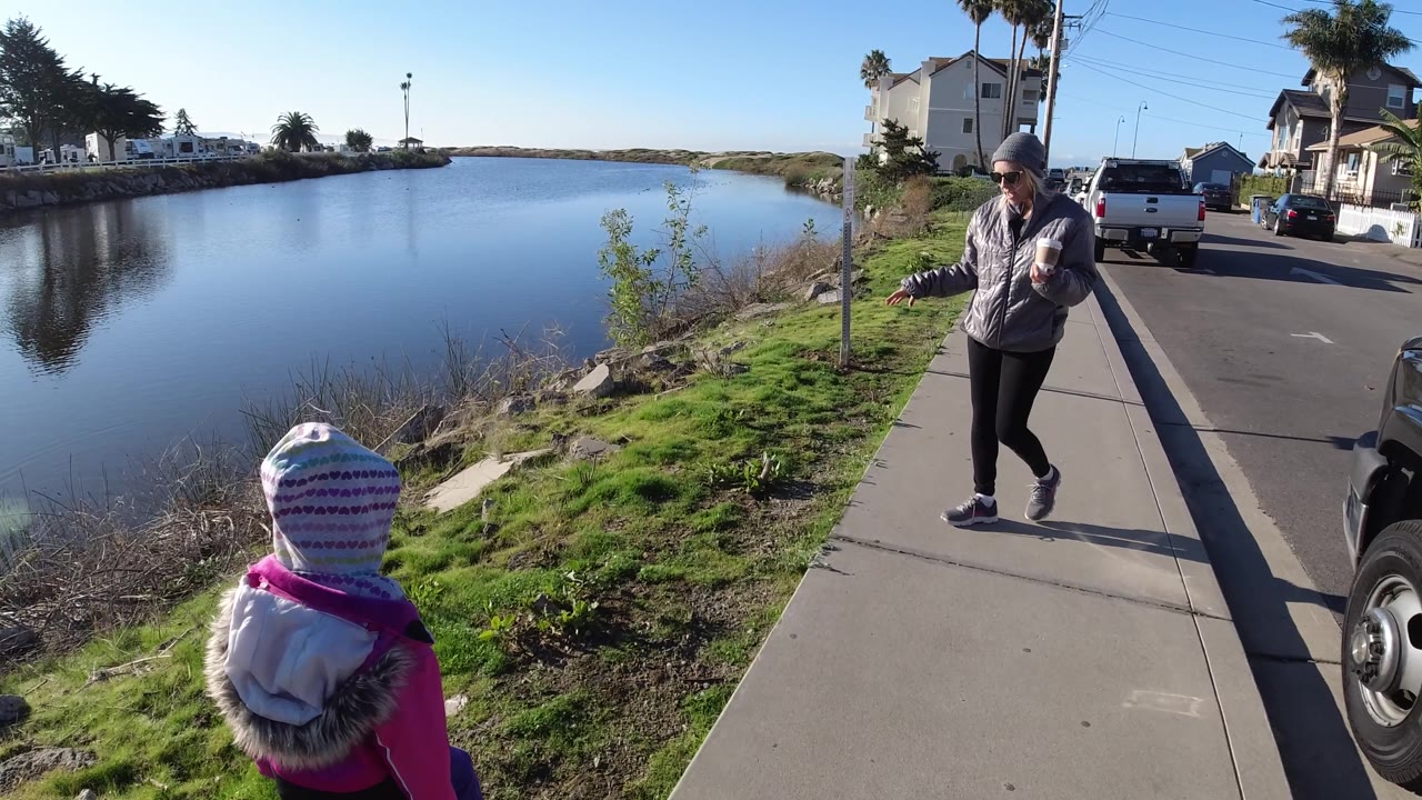 The woman faces back toward the camera while holding a drink, the child walks near the green bank, and the residential street and water remain visible in Pismo Beach.