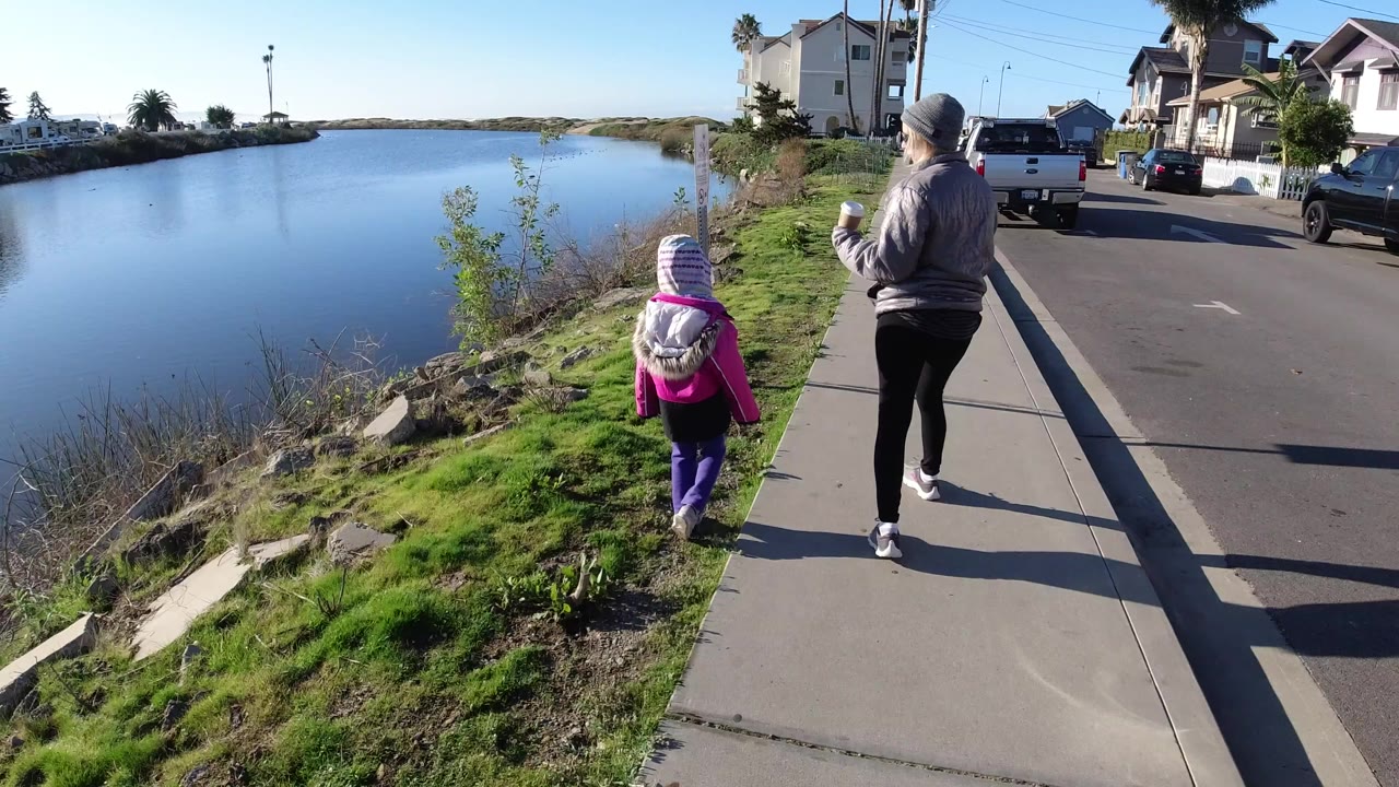 In Pismo Beach, California, a woman and child walk away from the camera on a sidewalk beside calm water, grass, parked cars, and residential houses.