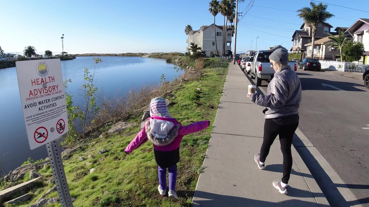 In Pismo Beach, a small child in pink walks beside an older woman on a sidewalk next to calm water, with a health advisory sign in the foreground.