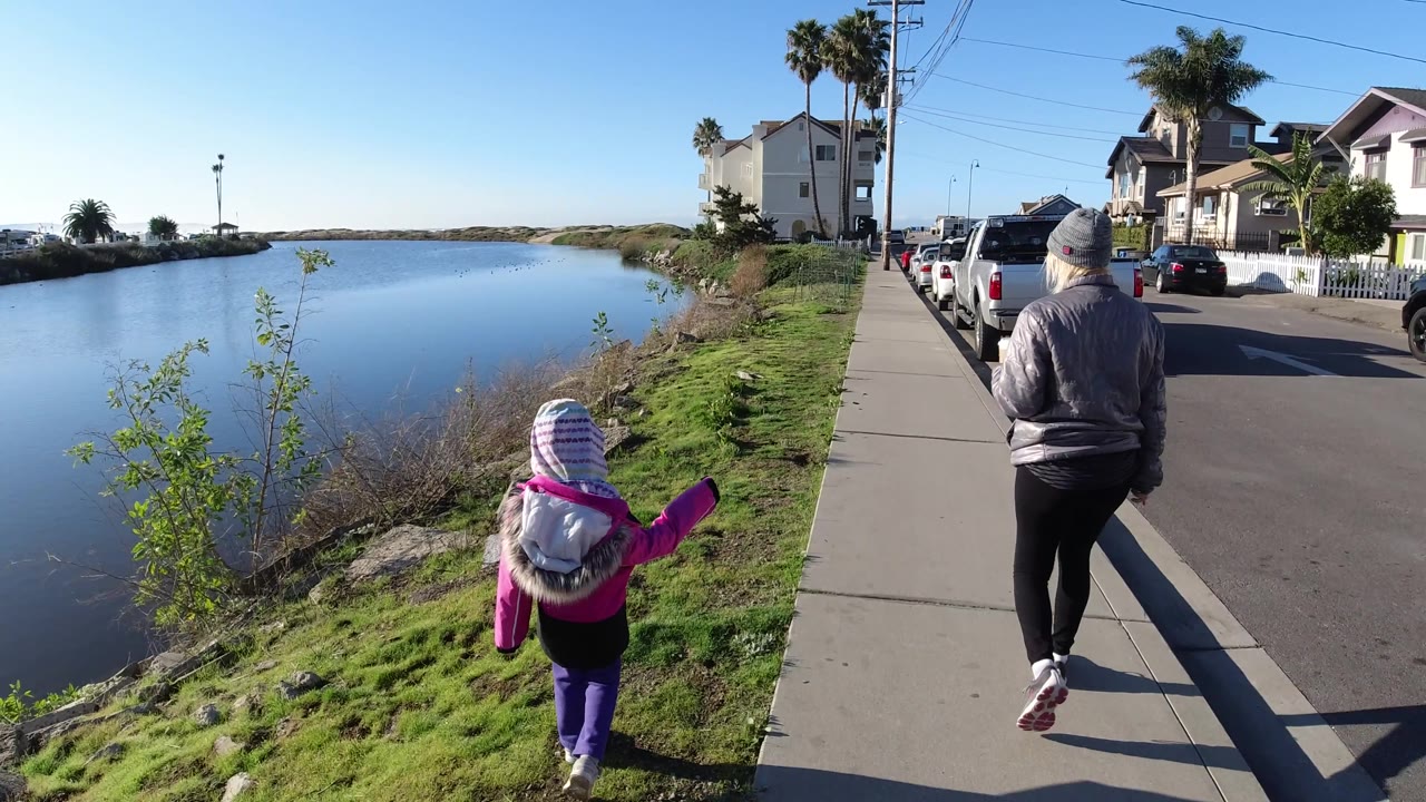 In Pismo Beach, the child moves ahead near the grass by the water while the older woman walks on the sidewalk beside a line of parked vehicles.