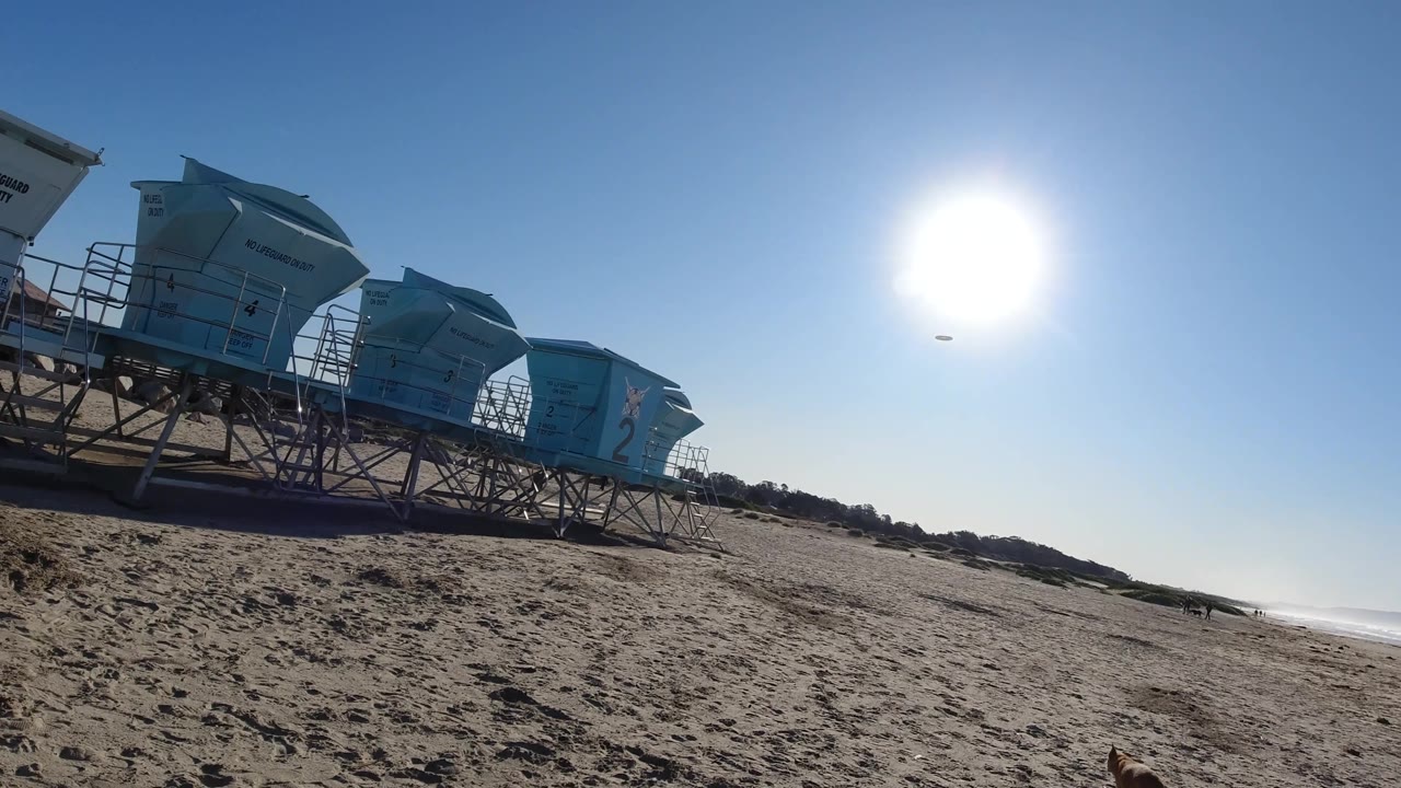 At Pismo Beach, blue lifeguard towers sit on raised supports at the left while the bright sun hangs over wide sand, distant surf, and no visible people.