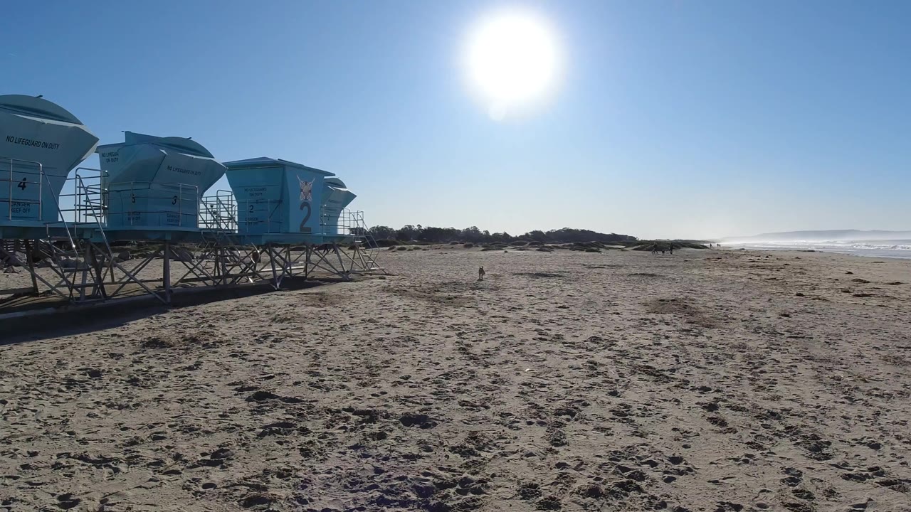 Pismo Beach appears from a similar wide angle with the lifeguard towers more centered, textured sand in the foreground, low surf at right, strong sun glare, and no visible people.