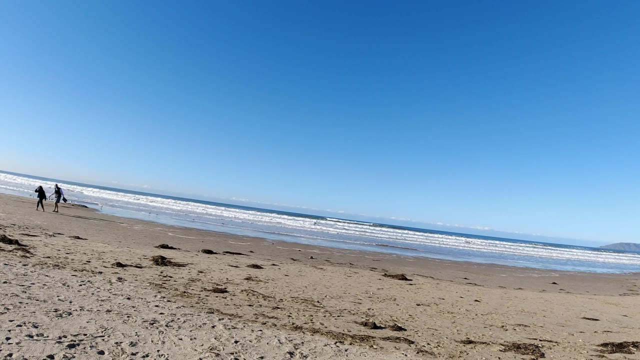 Two people walk along a wide beach with waves rolling behind them.