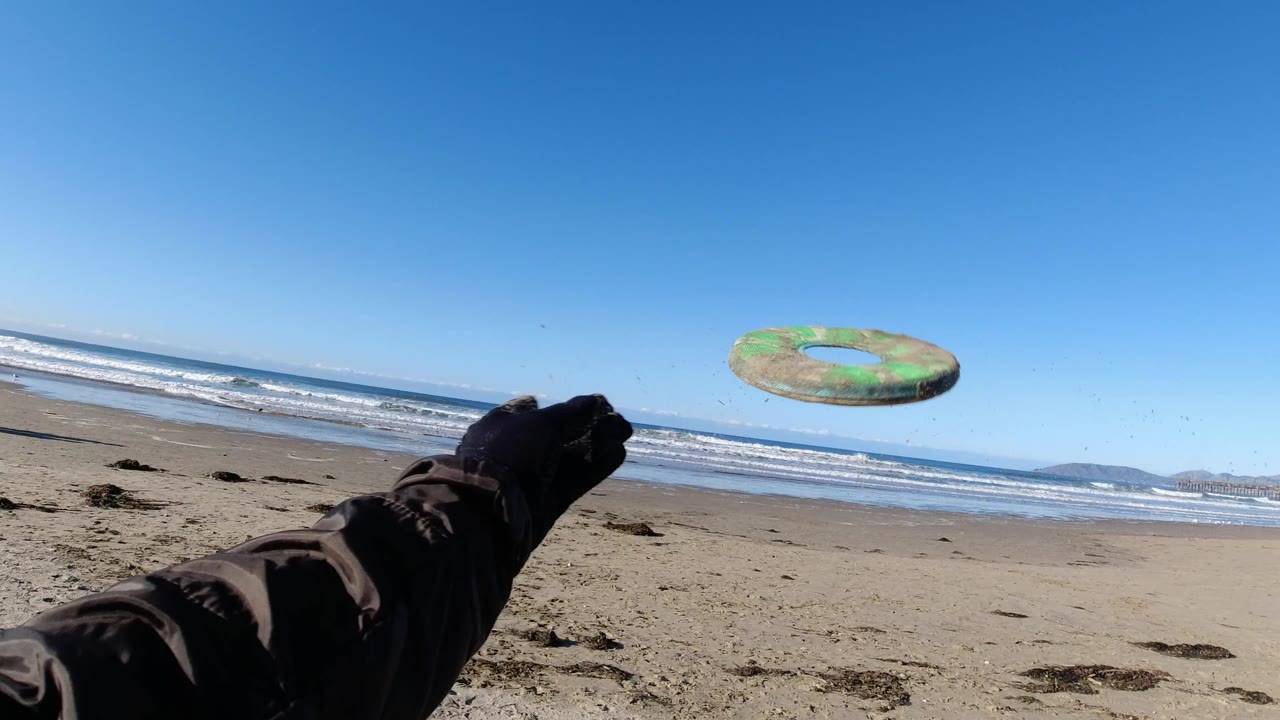 At Pismo Beach, a gloved foreground hand releases a green frisbee over sand, with blue sky, breaking waves, and low landforms in the distance.