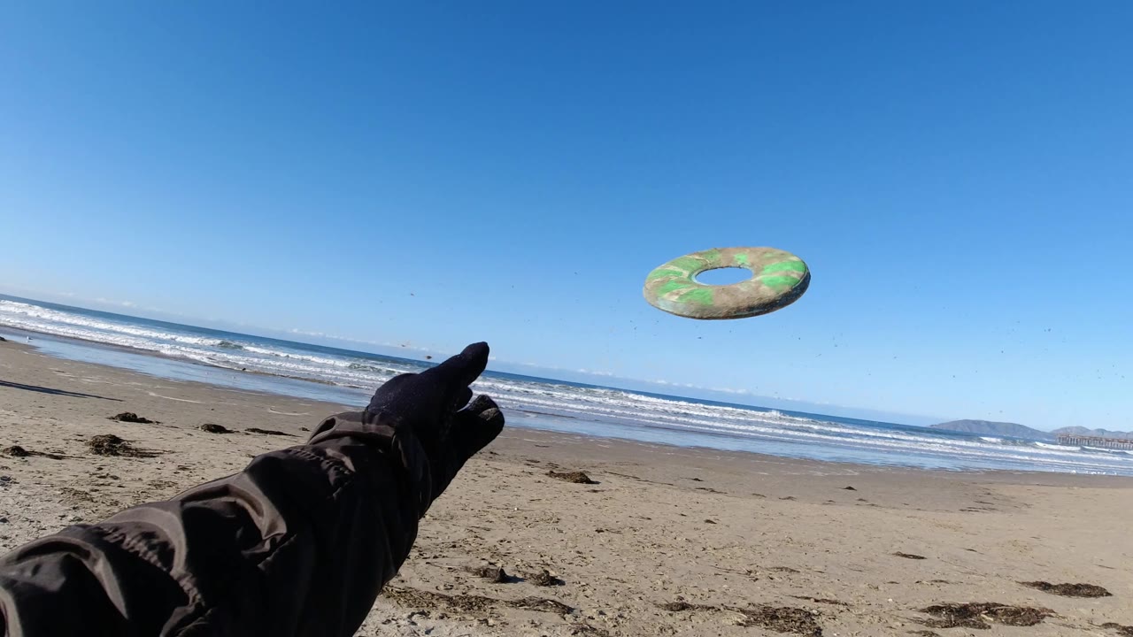 At Pismo Beach, the green frisbee is airborne above the shoreline while a gloved hand remains in the foreground and surf lines stretch across the background.