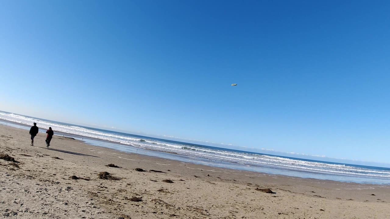 At Pismo Beach, two distant people walk along the sand while a small frisbee appears high against the blue sky above the ocean horizon.