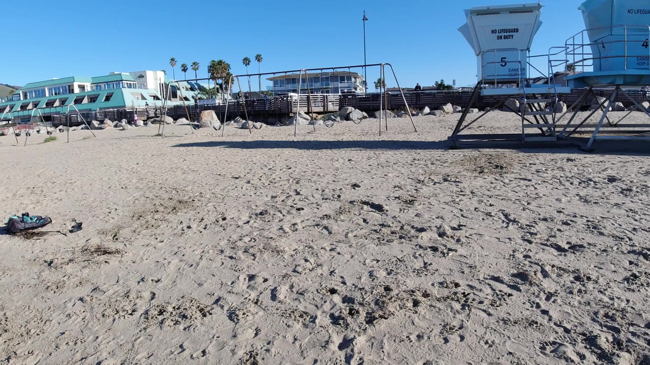 Wide Pismo Beach frame with pale sand in the foreground, a frisbee lying at lower left, beachfront buildings and palm trees behind it, a lifeguard tower at right, and no visible people.