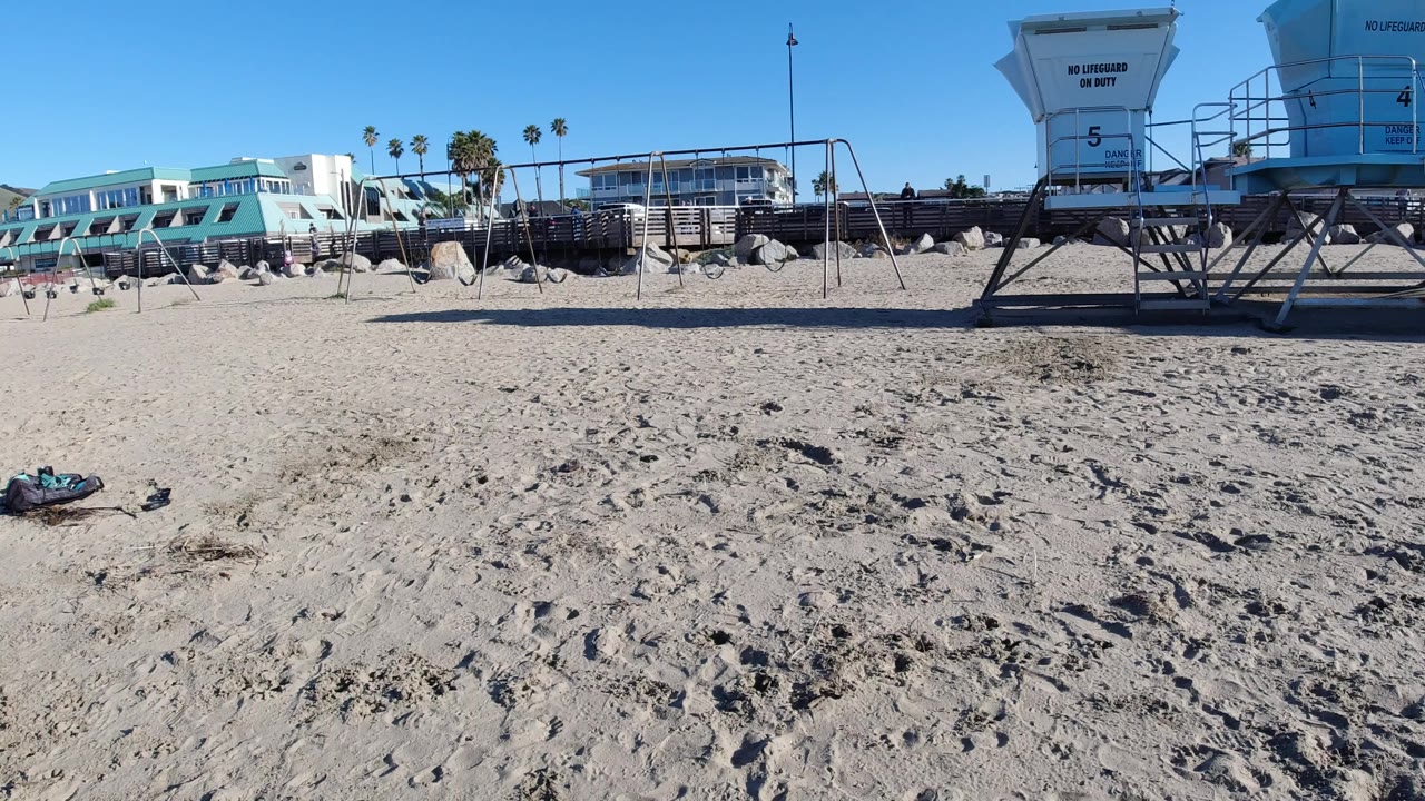 Pismo Beach view one second in, showing the frisbee low in frame, textured sand across the foreground, a lifeguard tower and swing frame on the right, coastal buildings in back, and no visible people.
