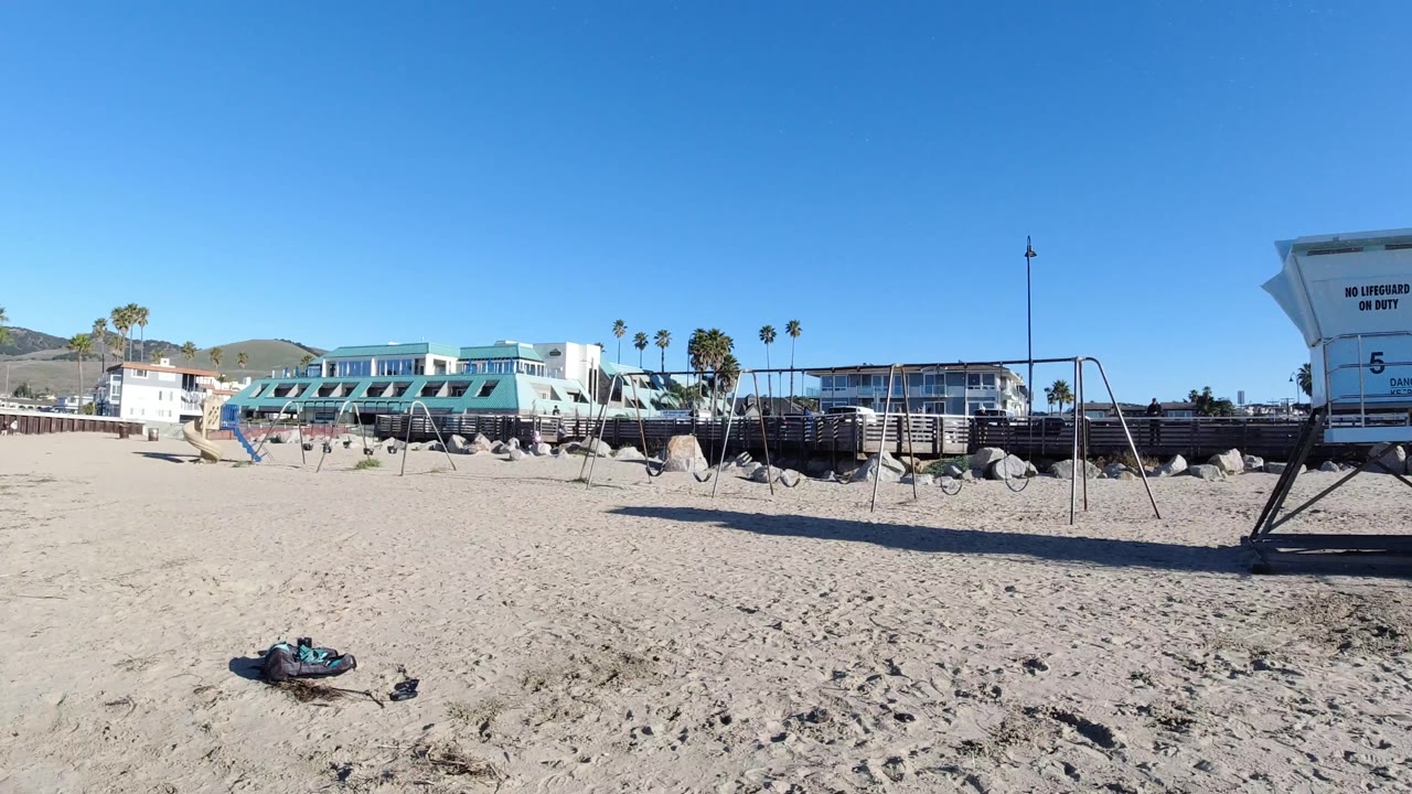 Wide Pismo Beach frame with the frisbee lower center-left, open sand leading to beachfront buildings and palm trees, the lifeguard tower cropped at the right edge, and no visible people.