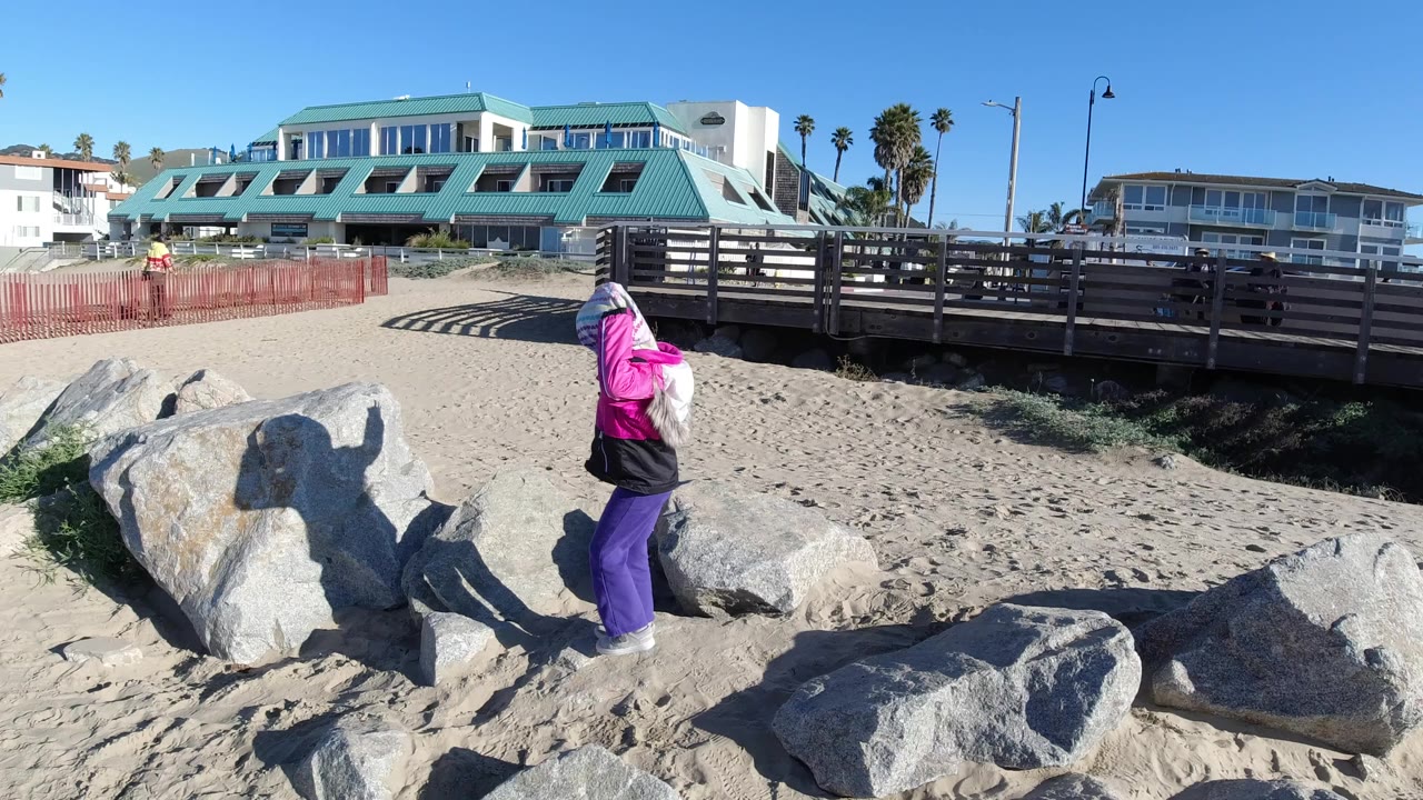 A child in a pink jacket and purple pants stands on large rocks at Pismo Beach, with sand, a wooden railing, palms, and low buildings behind them.