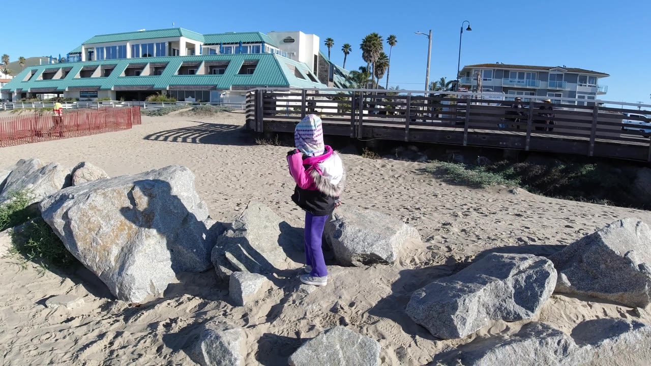 A rear view shows the child on the rocks at Pismo Beach, with shadows on the sand, a raised wooden rail, palm trees, and coastal buildings behind.