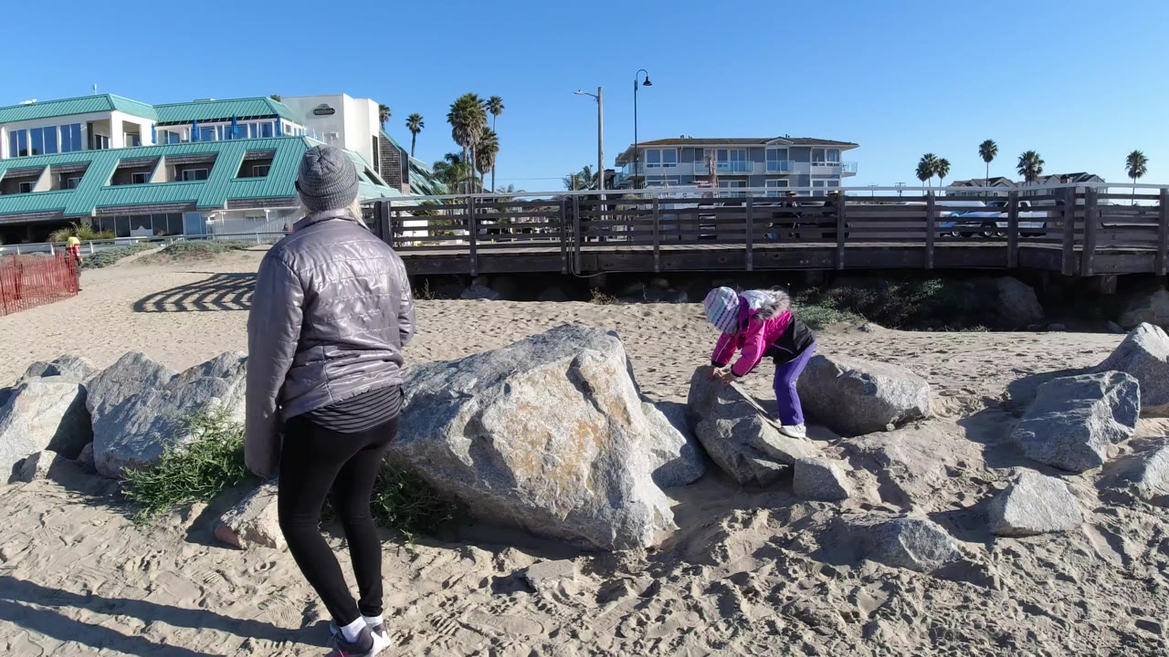 Adult watches as a child climbs over rocks on a sunny beach.