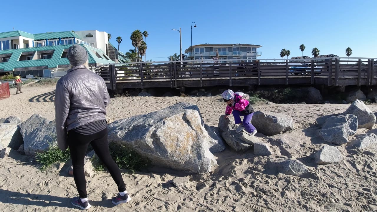 At Pismo Beach, the child shifts farther across the rocks in a pink jacket and purple pants as the adult stands with their back to camera, framed by sand, boulders, a boardwalk, and teal-roofed buildings.