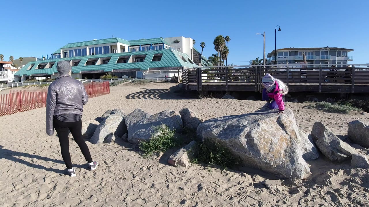 At Pismo Beach, an adult in a gray jacket stands with their back to camera while a child in bright clothing crouches near large rocks on the sand.