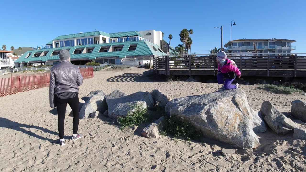 At Pismo Beach, the adult stands near the center foreground facing the rocks while the child stands higher on a boulder to the right.
