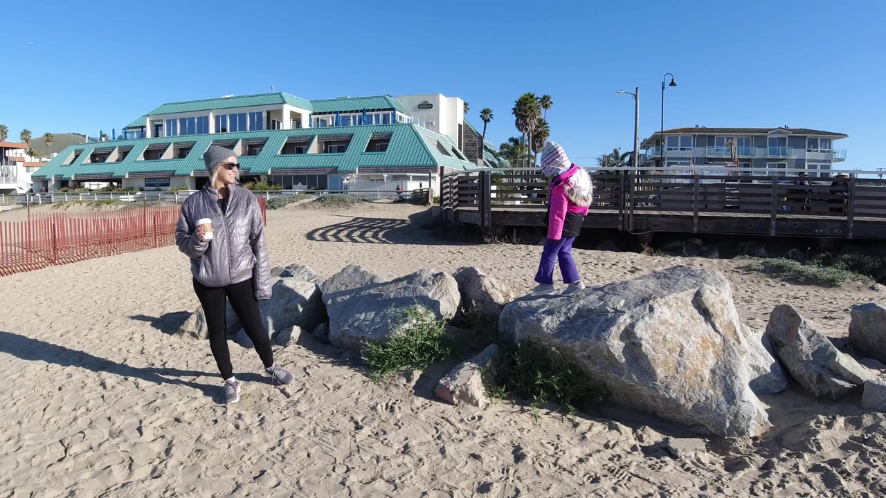 At Pismo Beach, an adult in a gray jacket stands on sand holding a cup while a girl in a pink jacket steps across large gray rocks near a wooden railing and beach buildings.