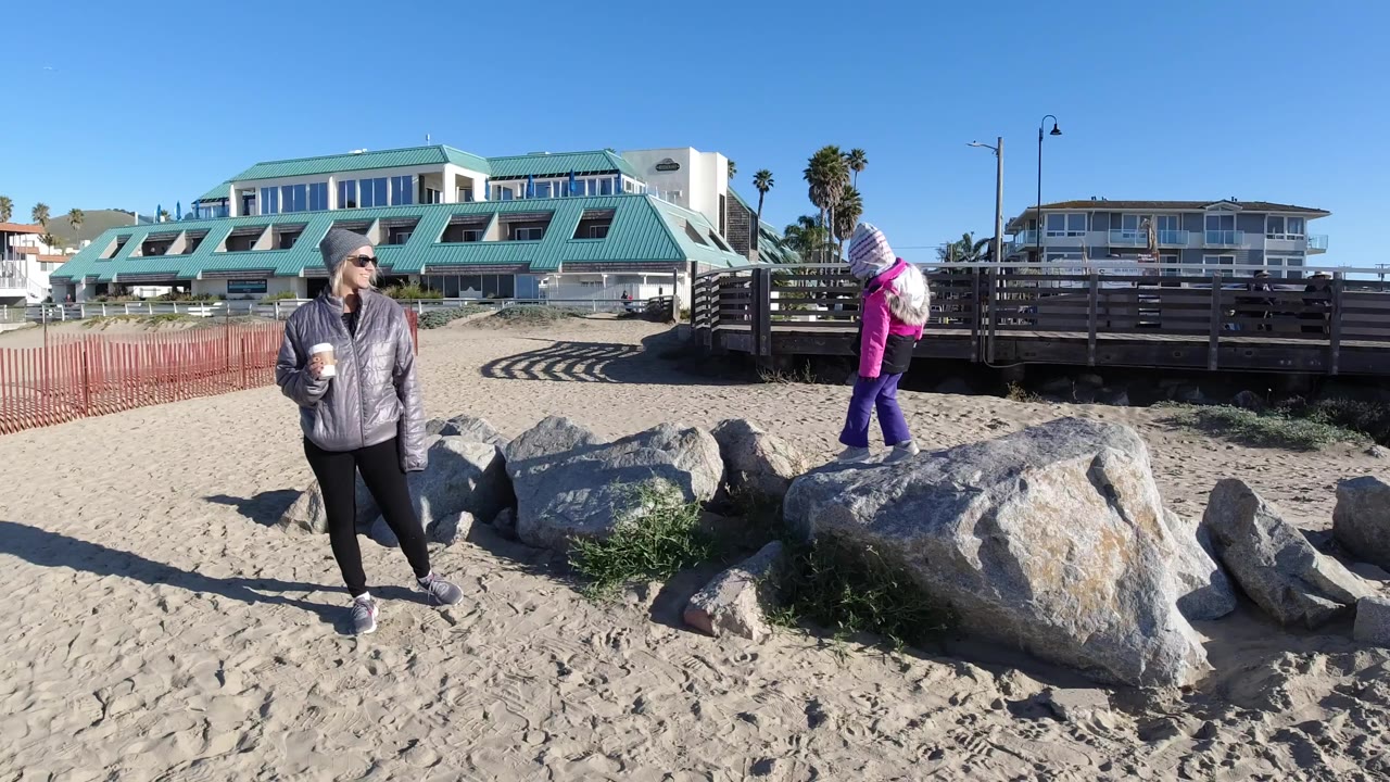 Girl in pink jacket stands on rocks while adult faces across the sand.