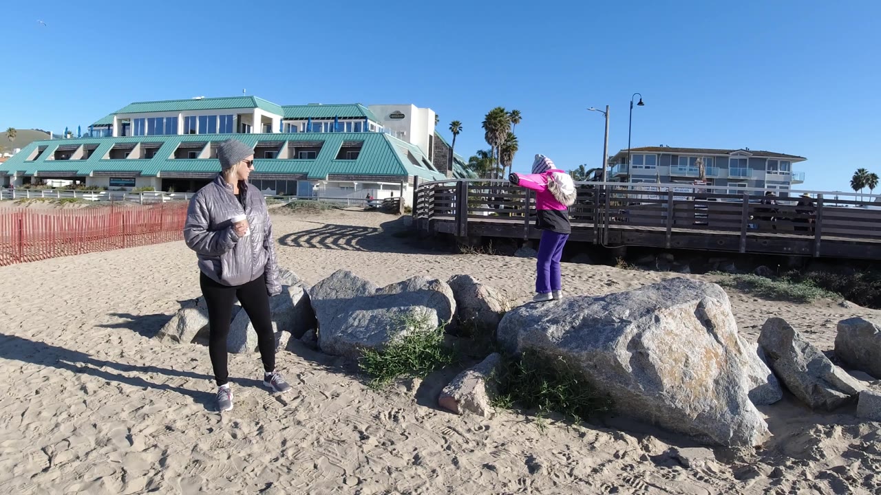 At Pismo Beach, the adult in gray looks toward the girl in pink as she extends an arm near the wooden railing, with large foreground rocks and bright sun across the sand.