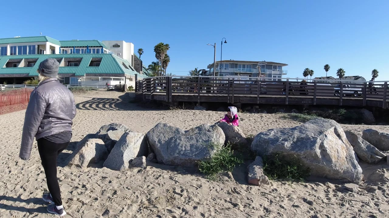 At 0:00 in Pismo Beach, California, a person in a gray jacket and beanie walks across sand toward large rocks where a person in pink sits near a wooden walkway.