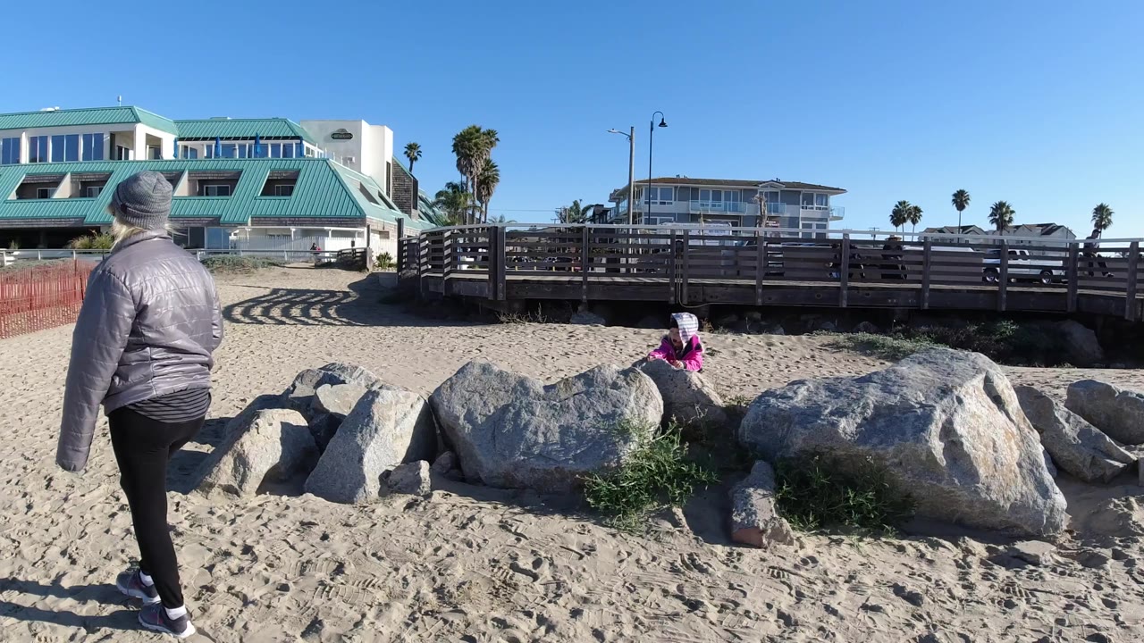 At 0:01 in Pismo Beach, California, the gray-jacketed person stands nearer the rocks while the person in pink remains seated or leaning on a stone below the walkway.