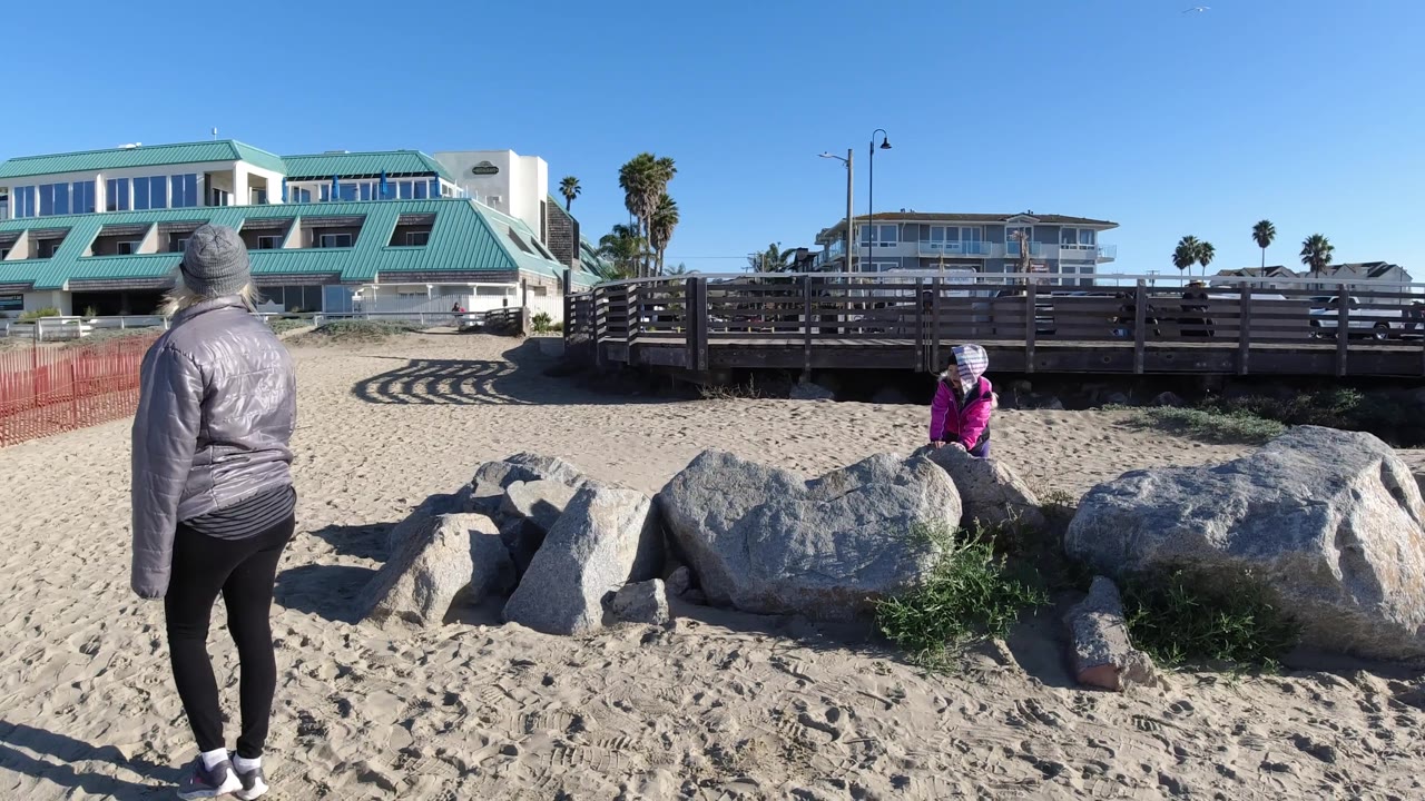 At 0:02 in Pismo Beach, California, the person in gray pauses near the center foreground, with the person in pink still positioned by the rock cluster and boardwalk.