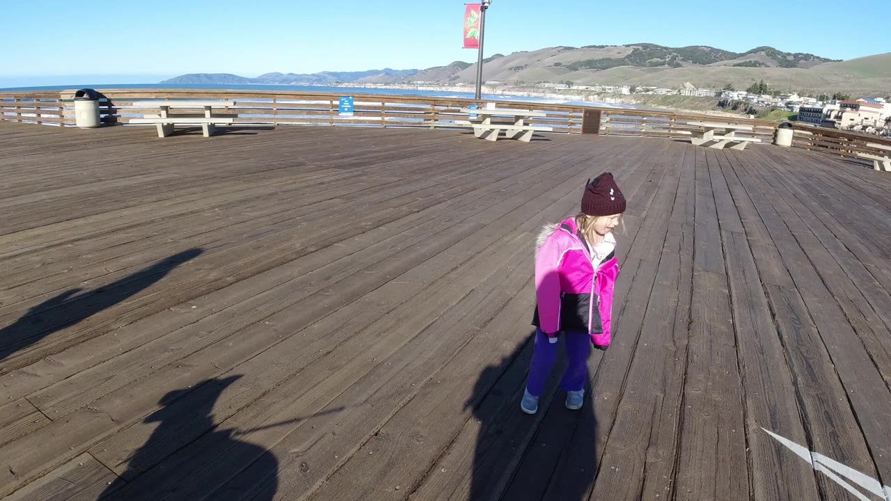 A child in a bright pink jacket and maroon knit hat stands on a wooden pier in Pismo Beach, California, with benches, railing, ocean, distant hills, and long shadows visible.