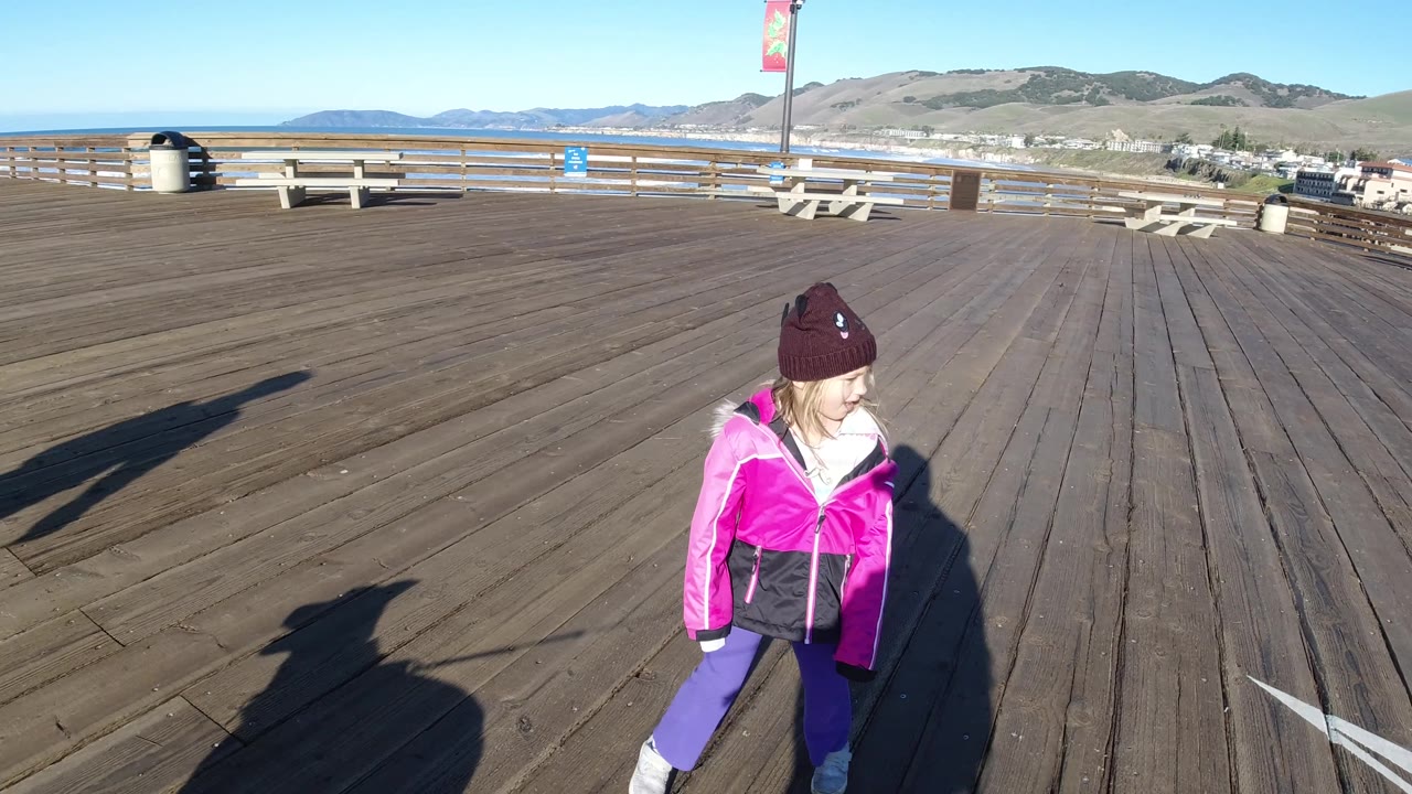 A closer right-side view shows the child walking across the Pismo Beach pier, with weathered wood planks, open railing, ocean, hills, and strong shadows from people outside the frame.