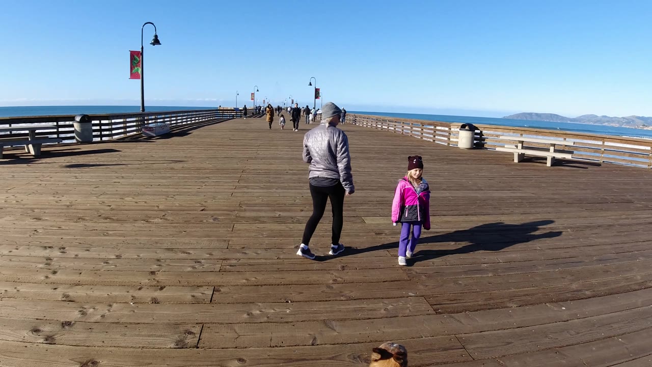 At Pismo Beach, California, the child in a pink jacket and purple pants walks near the center of the pier while the woman in gray moves slightly to the left, with open ocean, distant hills, benches, railings, and lamp posts visible.