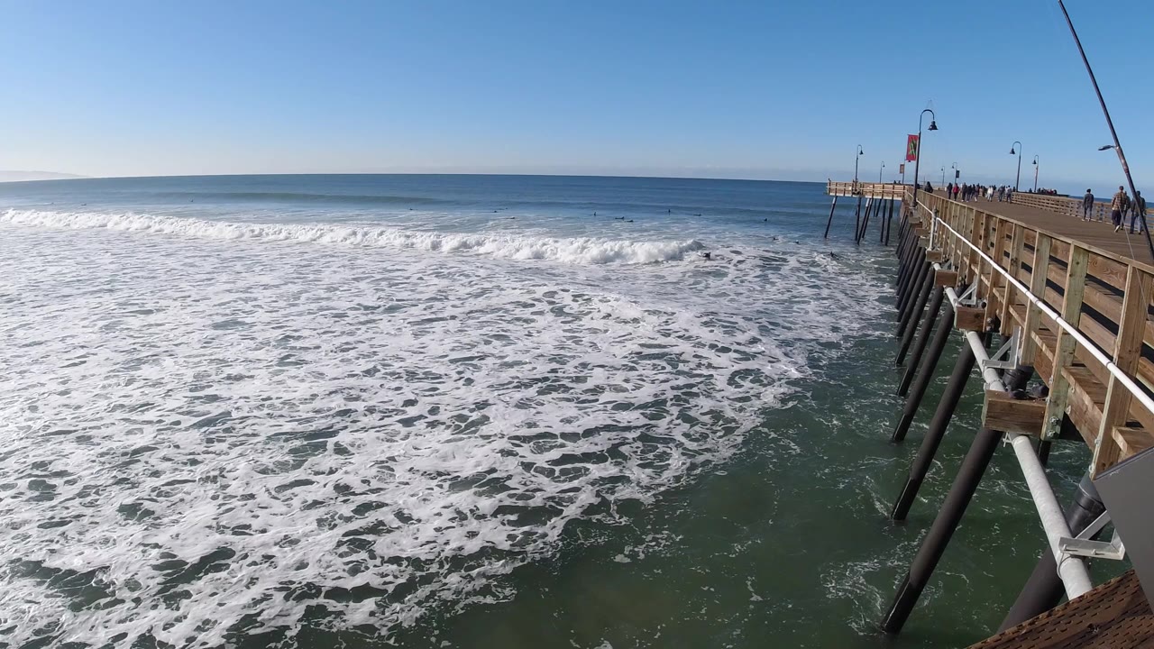 At 0:00, foamy surf fills the foreground at Pismo Beach, California, with a wooden pier and small distant pedestrians along the right side.