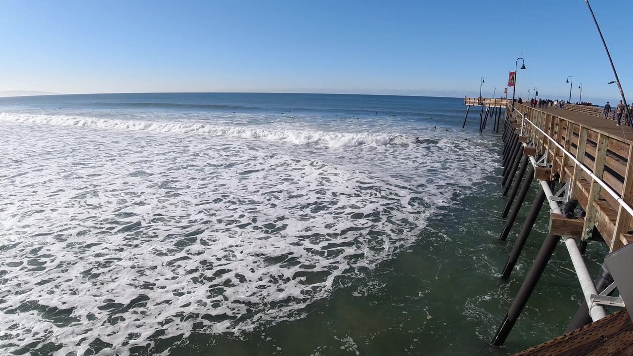 At 0:02, breaking white water stretches across the middle of the frame at Pismo Beach, California, with open ocean beyond and the pier cropped at right.