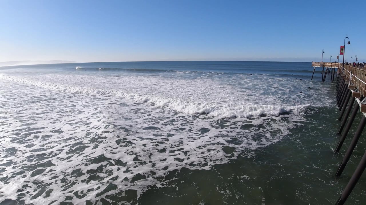 A breaking wave pushes toward the bridge posts at Pismo Beach, with open blue water beyond and small distant figures on the structure.