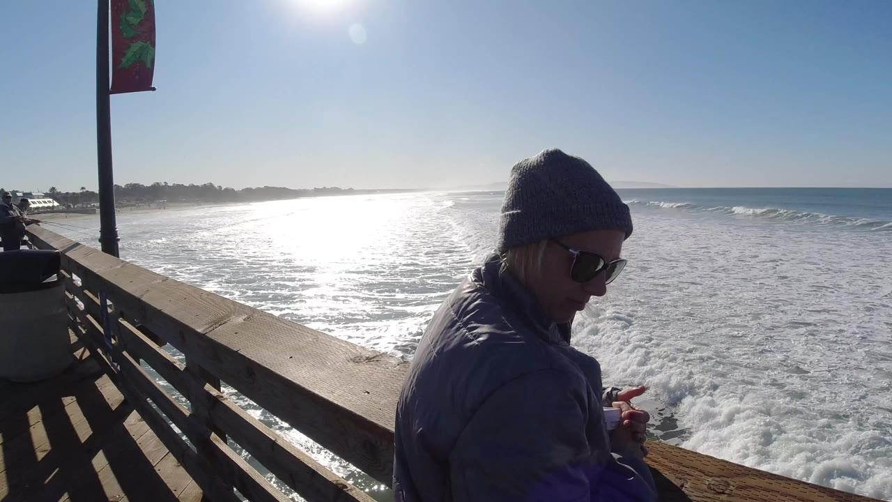At Pismo Beach, California, an adult in a knit beanie and sunglasses stands near a wooden platform rail while bright sun reflects across foamy ocean water.