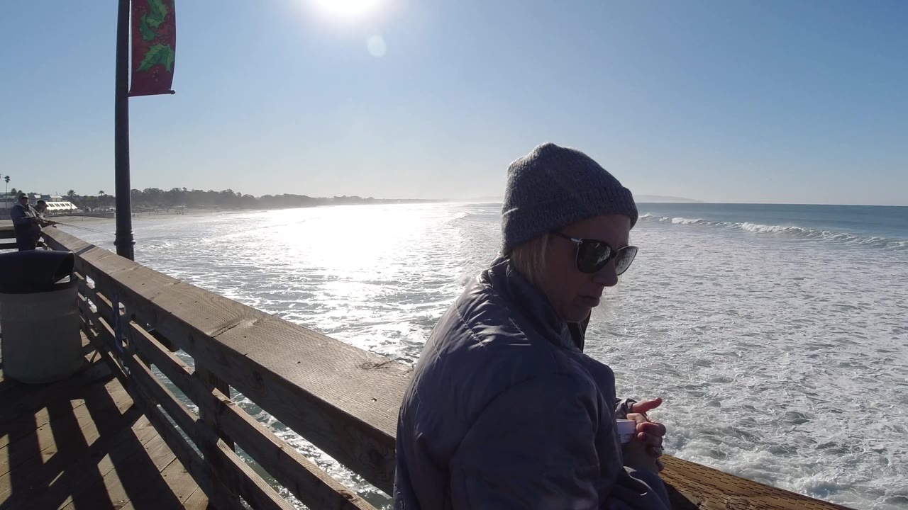 At Pismo Beach, California, the adult is cropped along the right edge while the wooden railing, trash can, and wide band of white ocean foam take more of the frame.