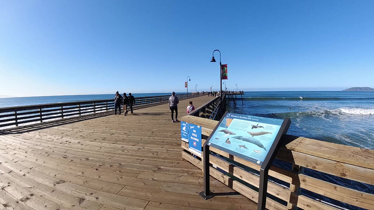Pismo Beach pier view with a wooden deck, railings, lamp posts, blue ocean, small surf, distant landform, foreground marine sign, and several small people far down the walkway.