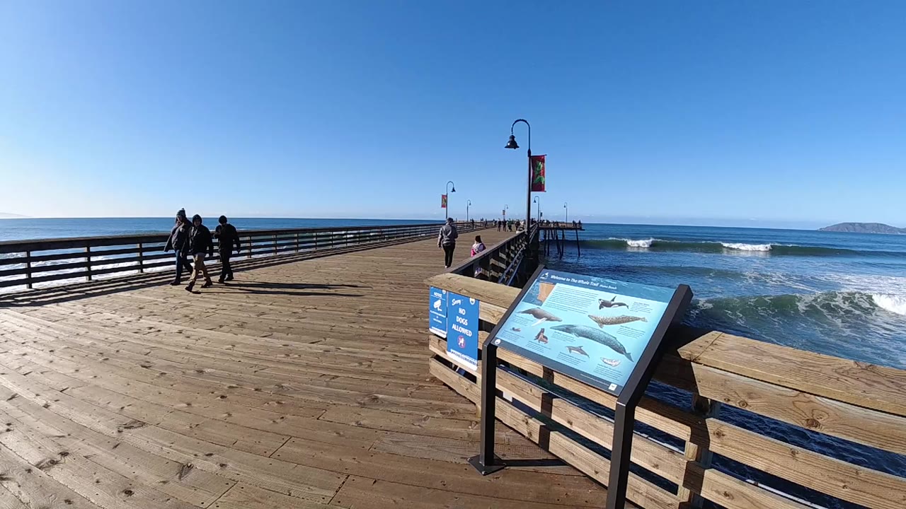 Wide view from a wooden pier in Pismo Beach, California, with an ocean information sign in the foreground, waves to the right, and several people walking along the railing.