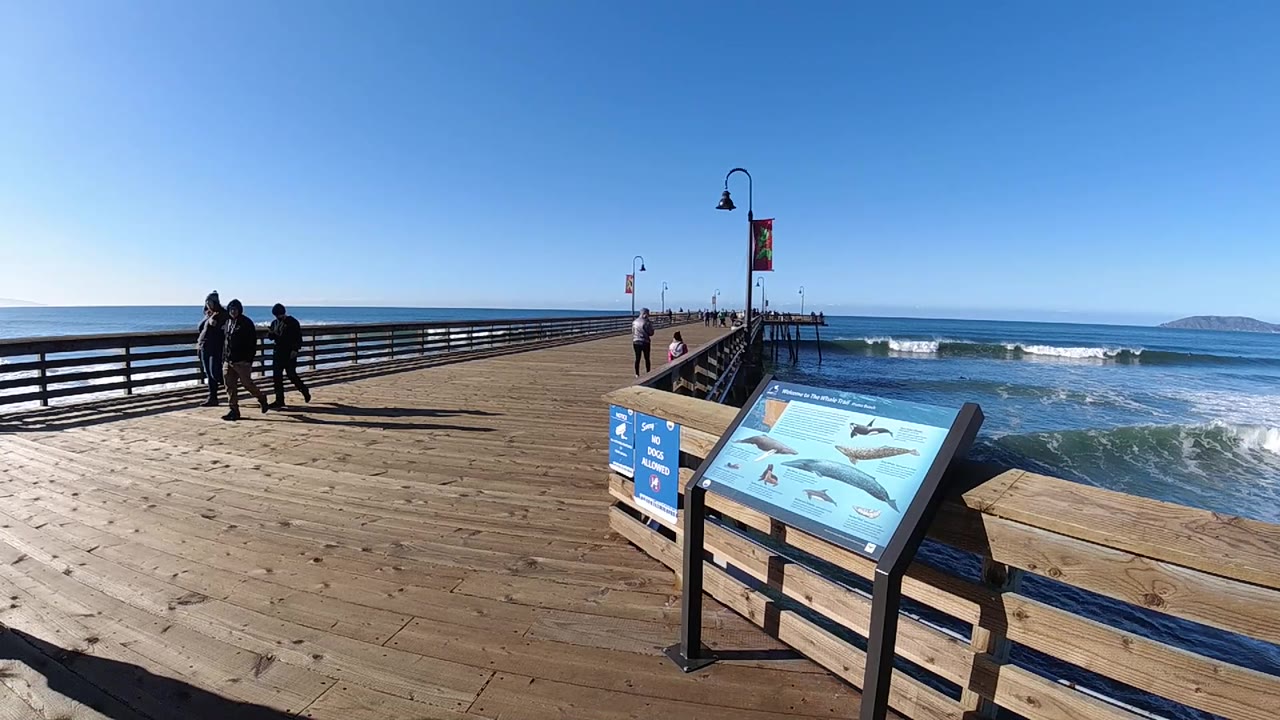 Wide Pismo Beach pier frame with the interpretive sign at lower right, people walking near the left railing, white surf rolling in, and a low landform on the horizon.