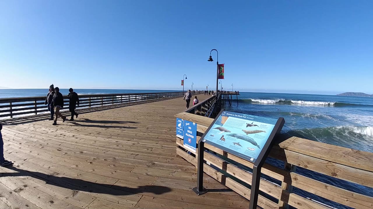 Wooden pier with walkers, railing, waves, and a foreground sign.