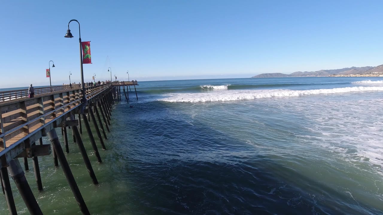 Pier railings and pilings beside breaking ocean waves under a clear sky.