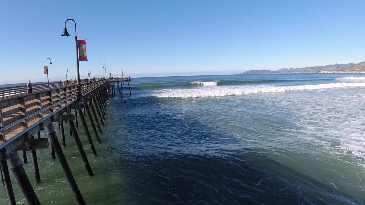 The Pismo Beach pier remains on the left with lamp posts and banners visible while a band of white surf spreads across the blue water toward the distant coastline.