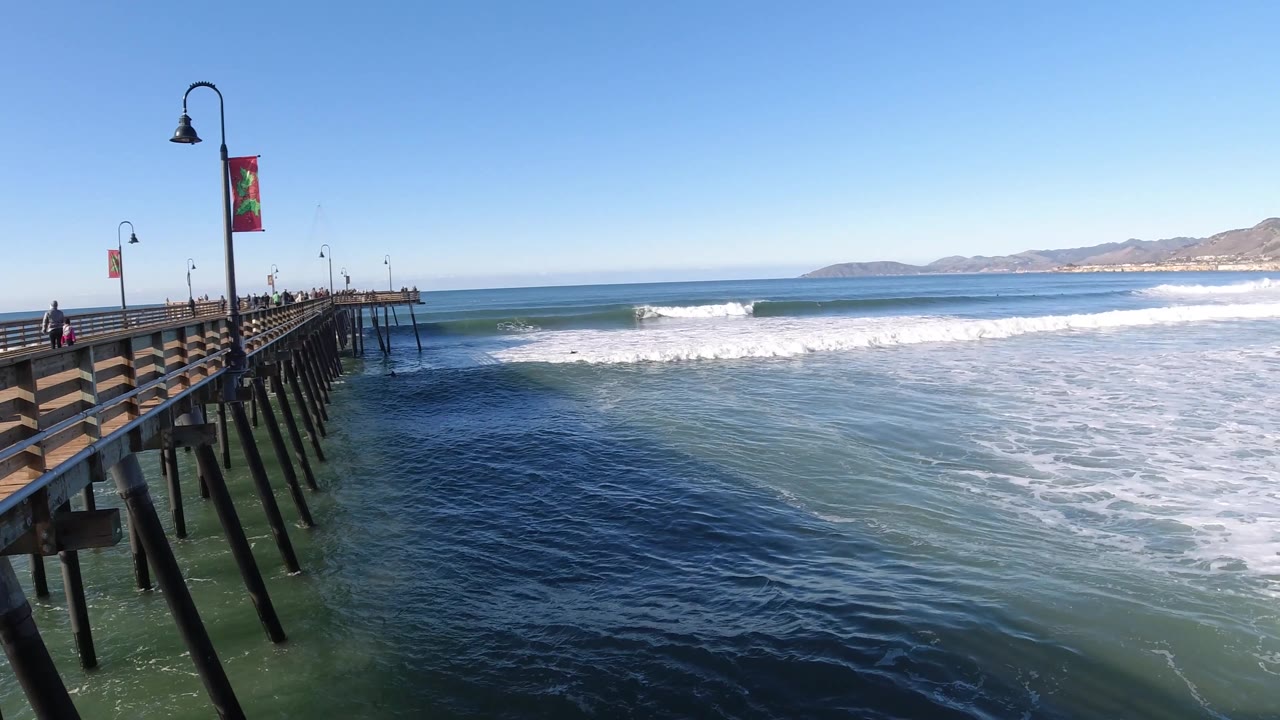 A slightly more rightward view of the Pismo Beach pier shows the railing, pilings, and lamp banners against dark blue water, white surf, and a pale coastline in the distance.