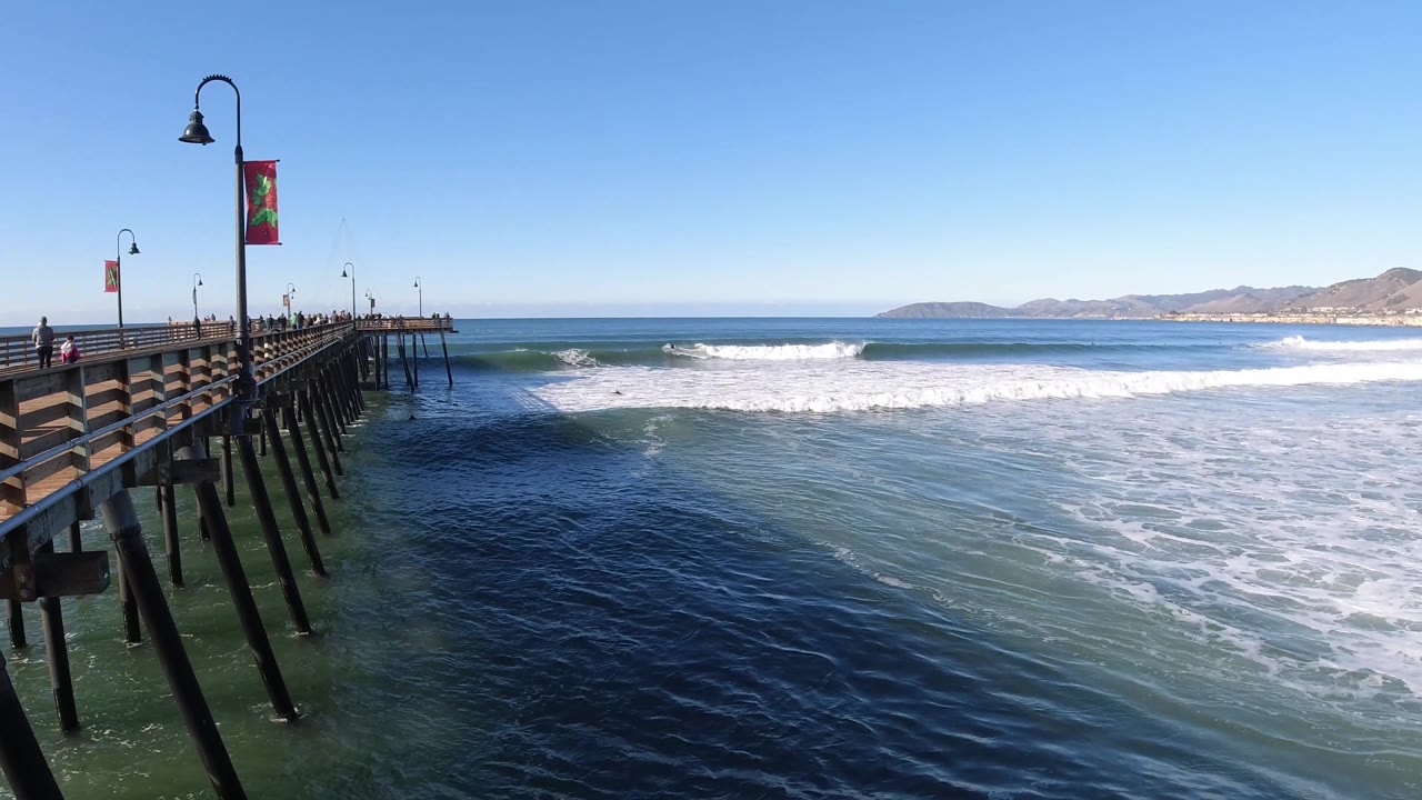 The Pismo Beach pier sits farther left in the frame as white foam widens over the water and the distant hills remain clear under a bright blue sky.