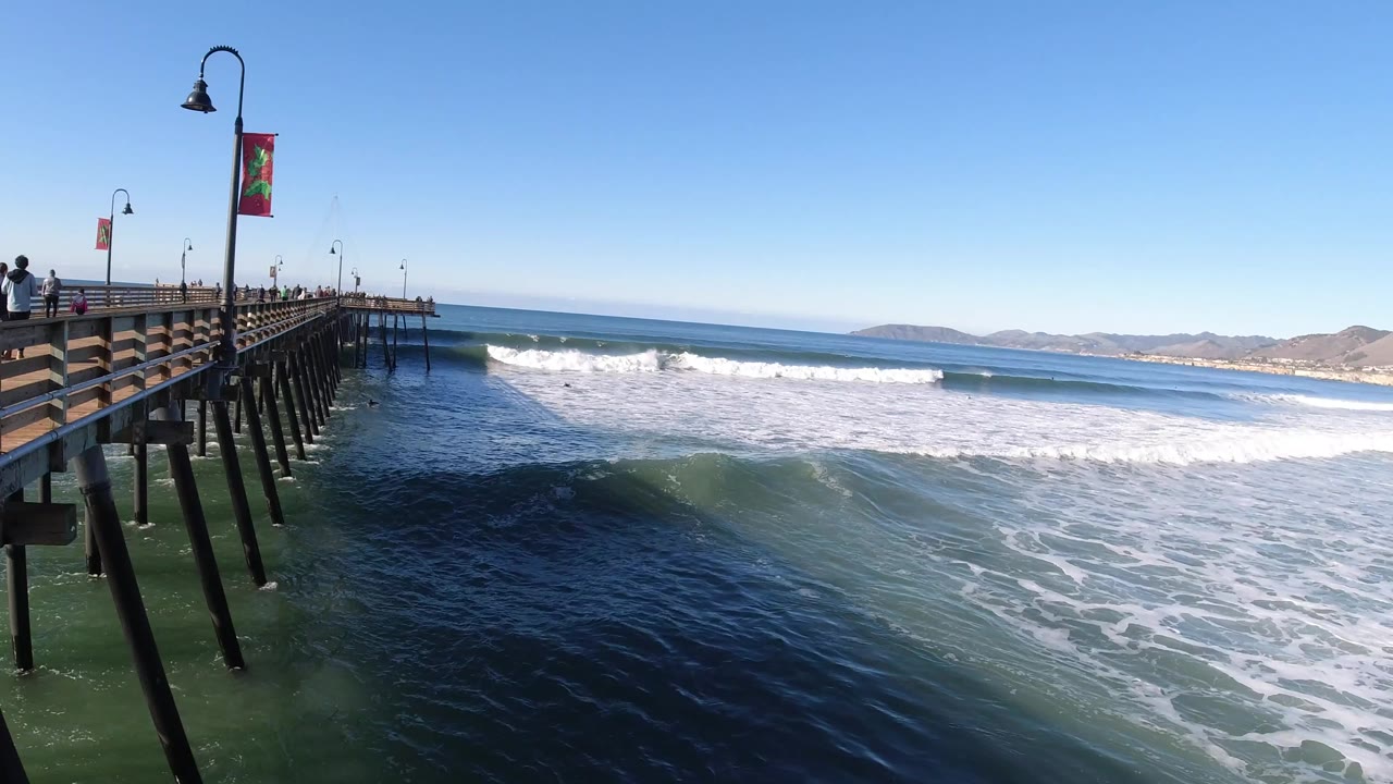 Pismo Beach ocean waves break beside a wooden pier with lamp posts and banners, with small distant people visible on the pier.
