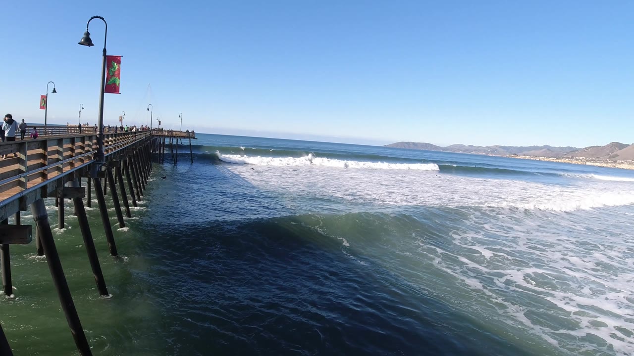 A darker wave line rises below the Pismo Beach pier, with white surf beyond it, low hills on the horizon, and small distant people visible near the railing.