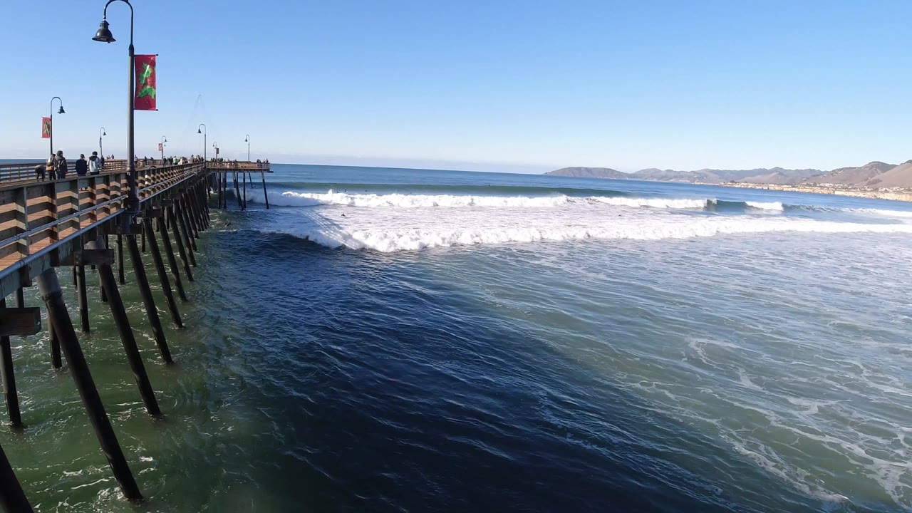 Pismo Beach pier lines the left side while white surf breaks across blue-green ocean and small people stand along the railing in clear daylight.