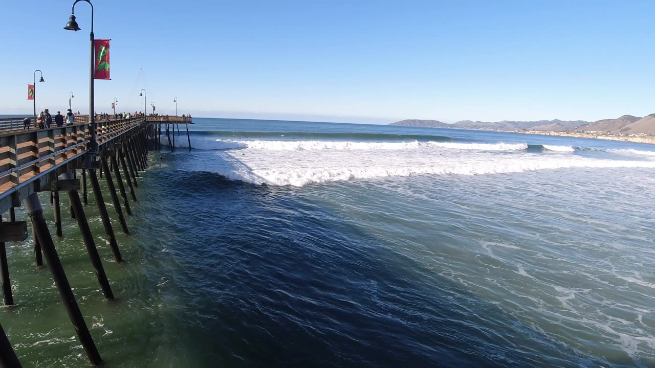 The Pismo Beach pier sits left of frame with small figures on the deck, while darker water in the foreground contrasts with bright foam across the surf zone.