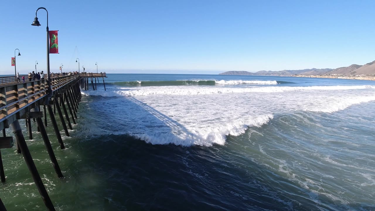 Pismo Beach pier remains on the left as a band of whitewater rolls across the center of the frame beneath a clear blue sky.