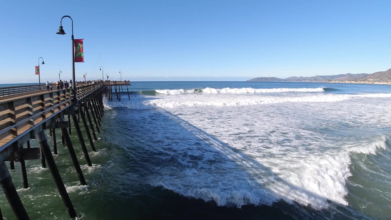 Ocean foam spreads below a pier with distant coastline beyond.