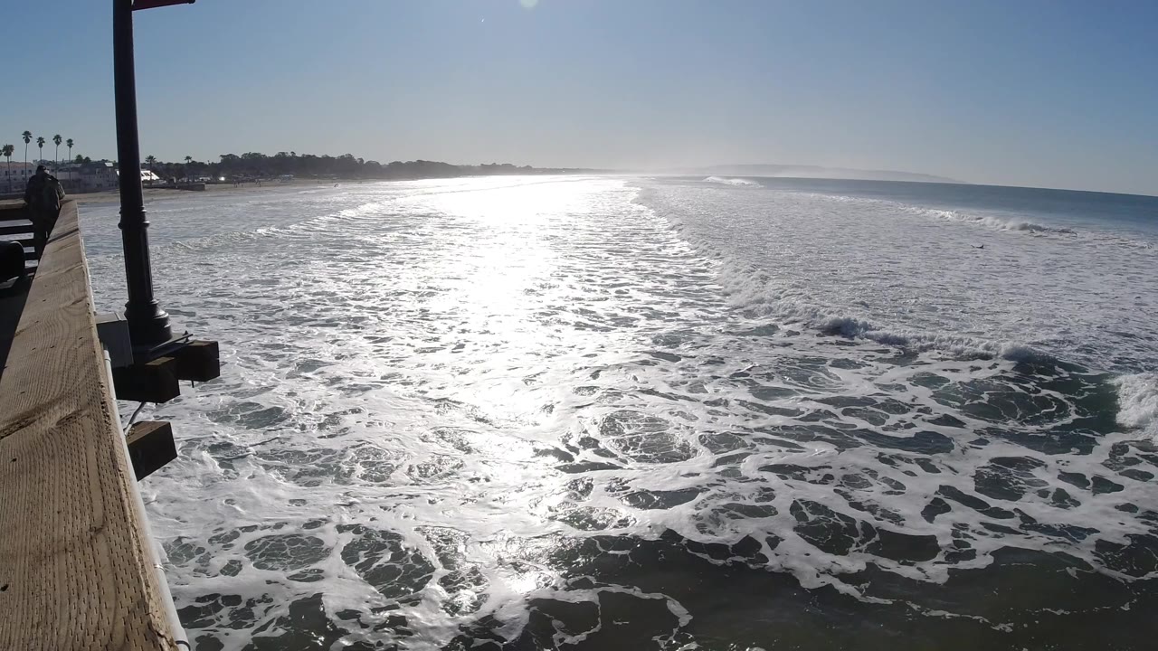 Bright sun glare spreads across breaking waves in Pismo Beach, California, with the pier edge and lampposts on the left and no visible people.