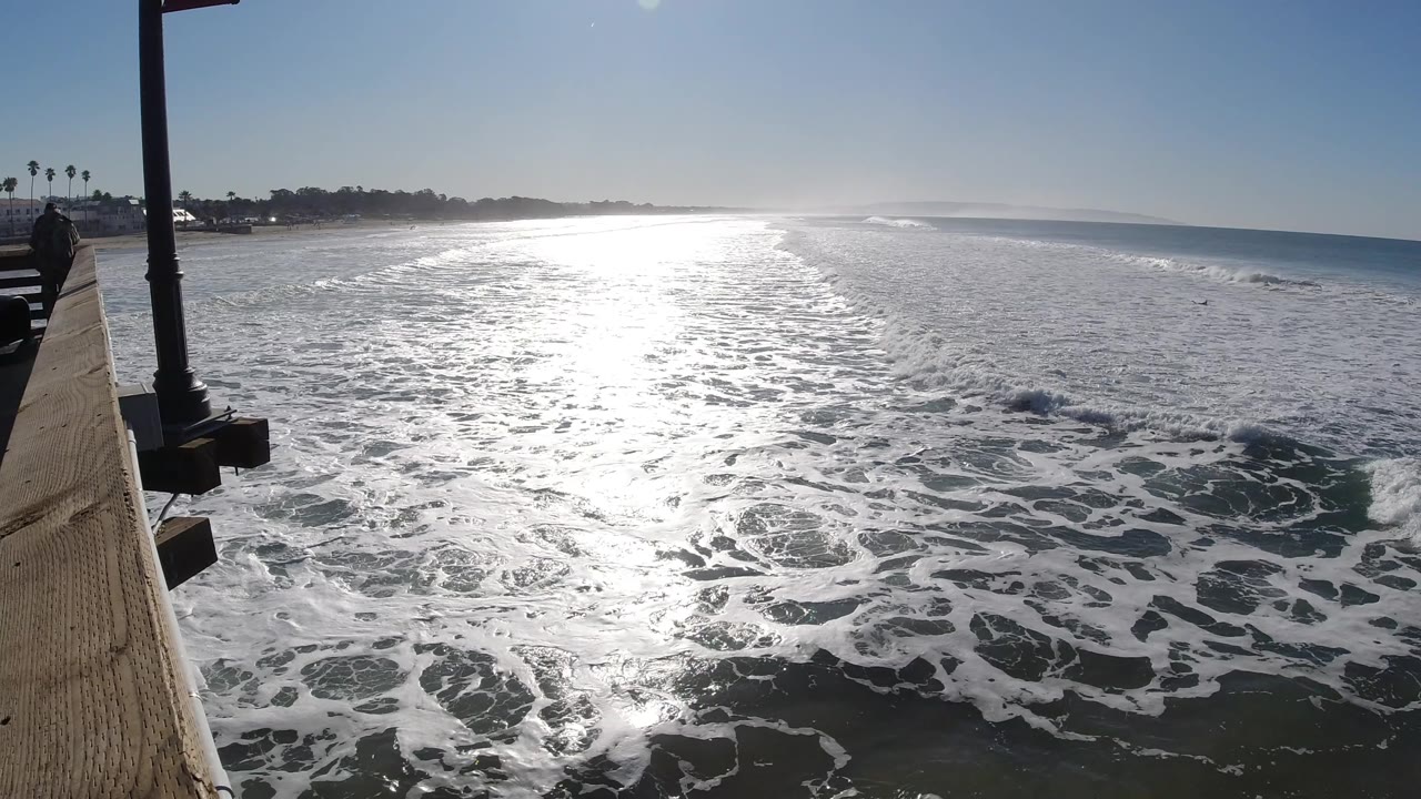 Foamy whitewater fills the foreground below a Pismo Beach pier-side view, with strong sunlight on the water and the horizon stretching rightward, no visible people.
