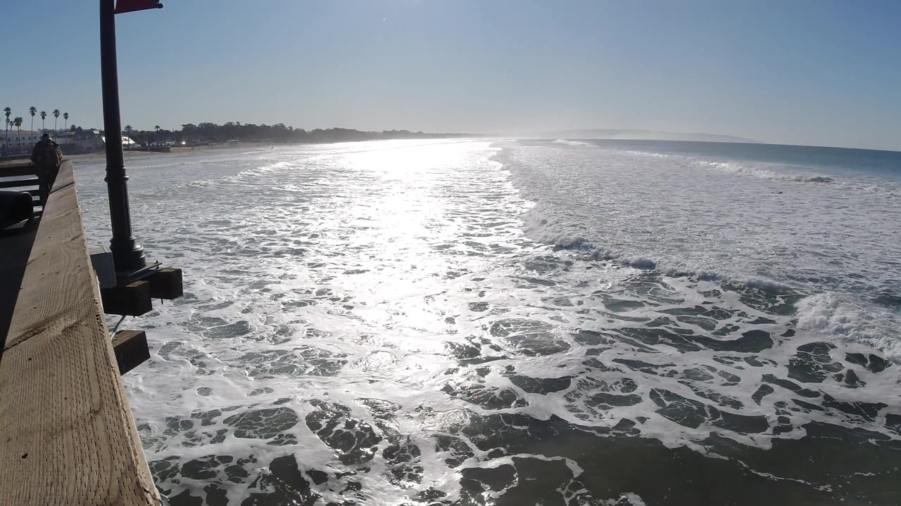 A wide Pismo Beach ocean frame shows white surf bands rolling toward the pier, a bright reflective path on the water, and no visible people.